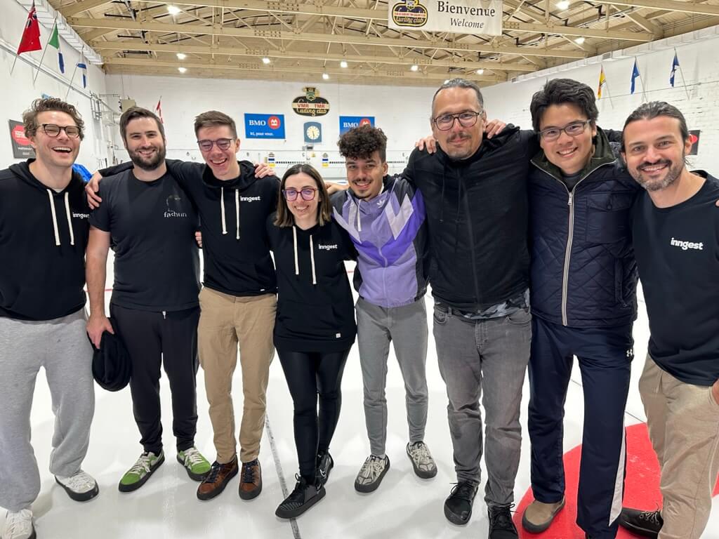 Eight happy people at a curling rink, standing arm to arm and laughing at a joke someone must have just told